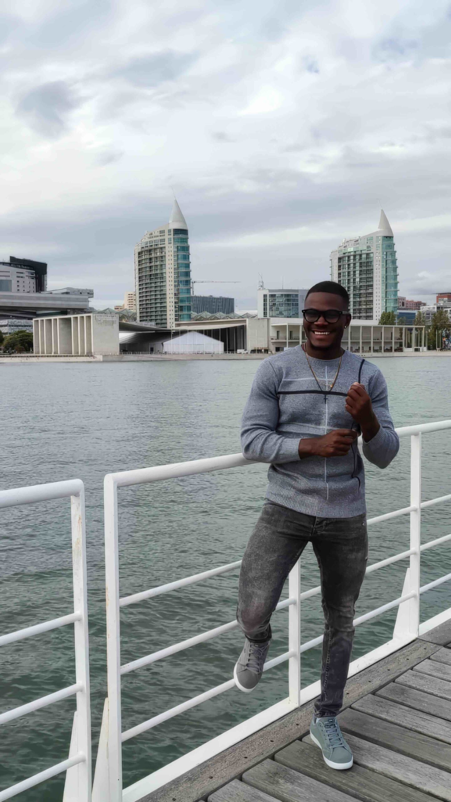 A casual portrait of Januário Luvemba standing on a riverside deck with a modern cityscape in the background. He is wearing a light grey jumper, dark jeans, and grey trainers, smiling warmly while posing confidently. The setting features contemporary architecture, calm water, and a cloudy sky, creating a relaxed yet urban atmosphere